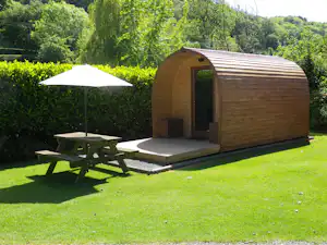 Holzfass-Hütte auf Campingplatz mit Terrasse, Liegefläche und Sonnenschirm, umgeben von Hecken und Bäumen.