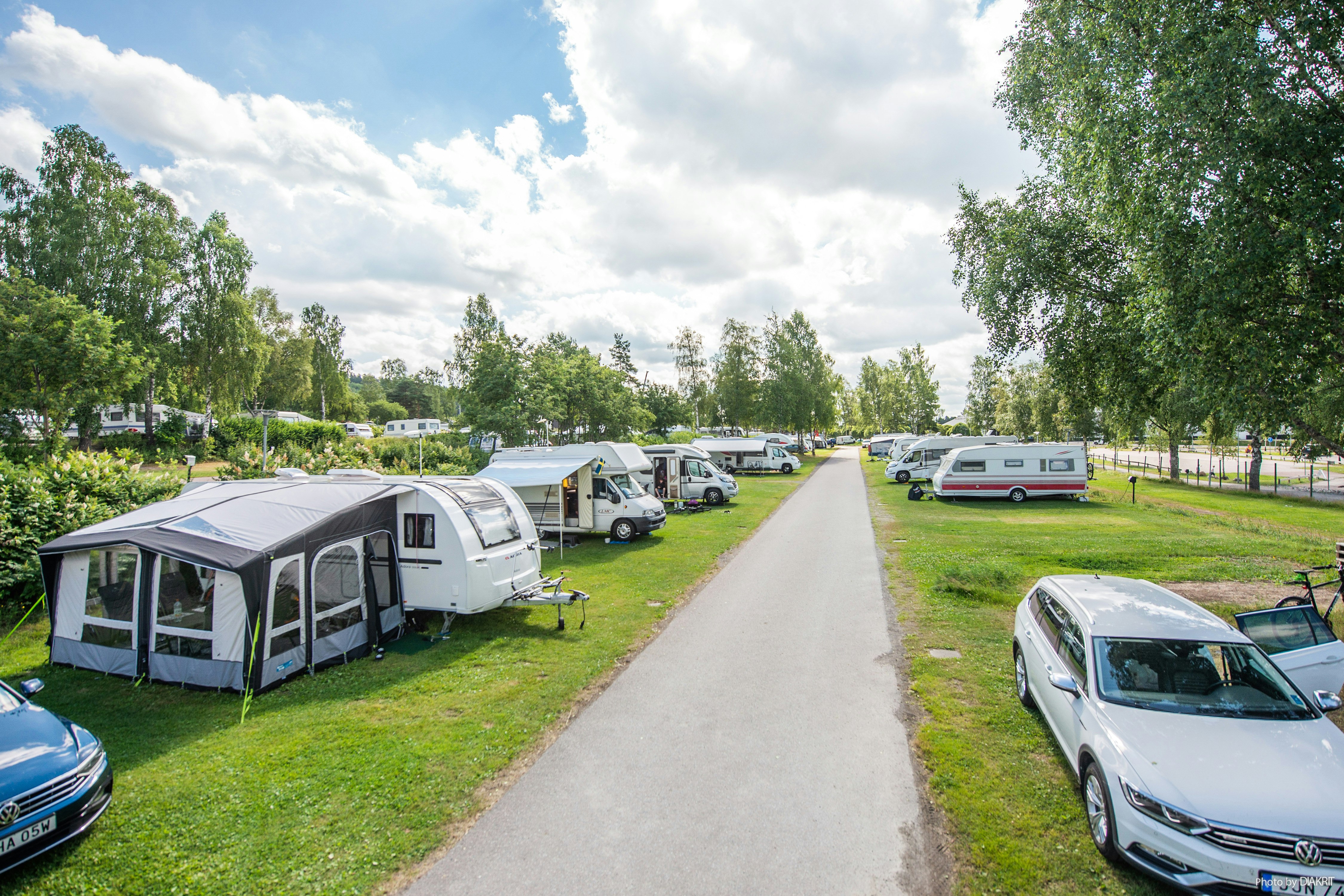 Wohnwagen und Zelte auf einem Campingplatz, dazwischen ein breiter Weg, geparkte Autos und Bäume entlang der Stellplätze.