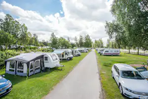 Wohnwagen und Zelte auf einem Campingplatz, dazwischen ein breiter Weg, geparkte Autos und Bäume entlang der Stellplätze.