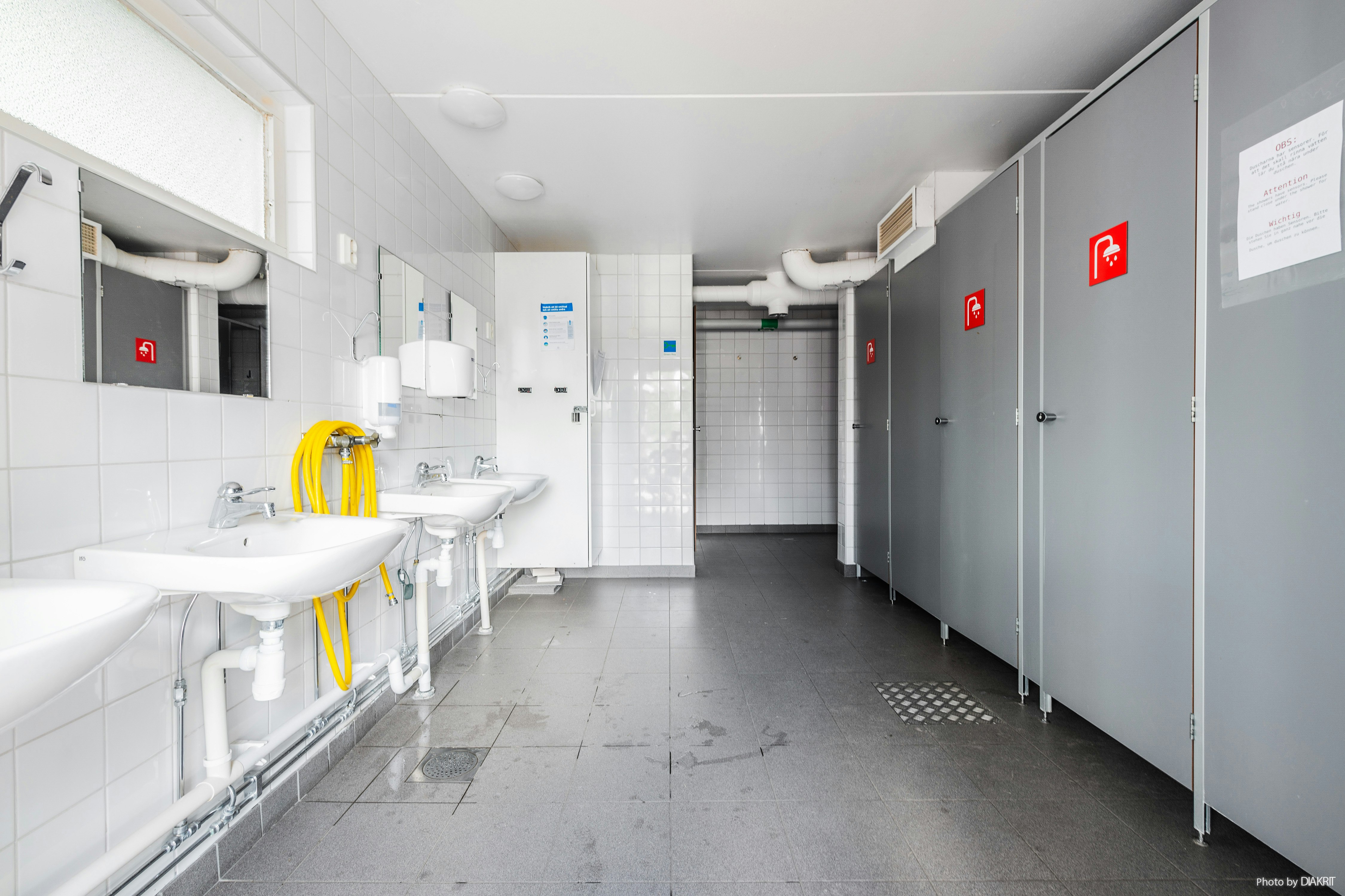 Helles Sanitärgebäude mit Waschbecken, Spiegeln und grauem Boden, daneben mehrere Türen und Schilder an den Wänden.