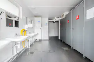 Helles Sanitärgebäude mit Waschbecken, Spiegeln und grauem Boden, daneben mehrere Türen und Schilder an den Wänden.