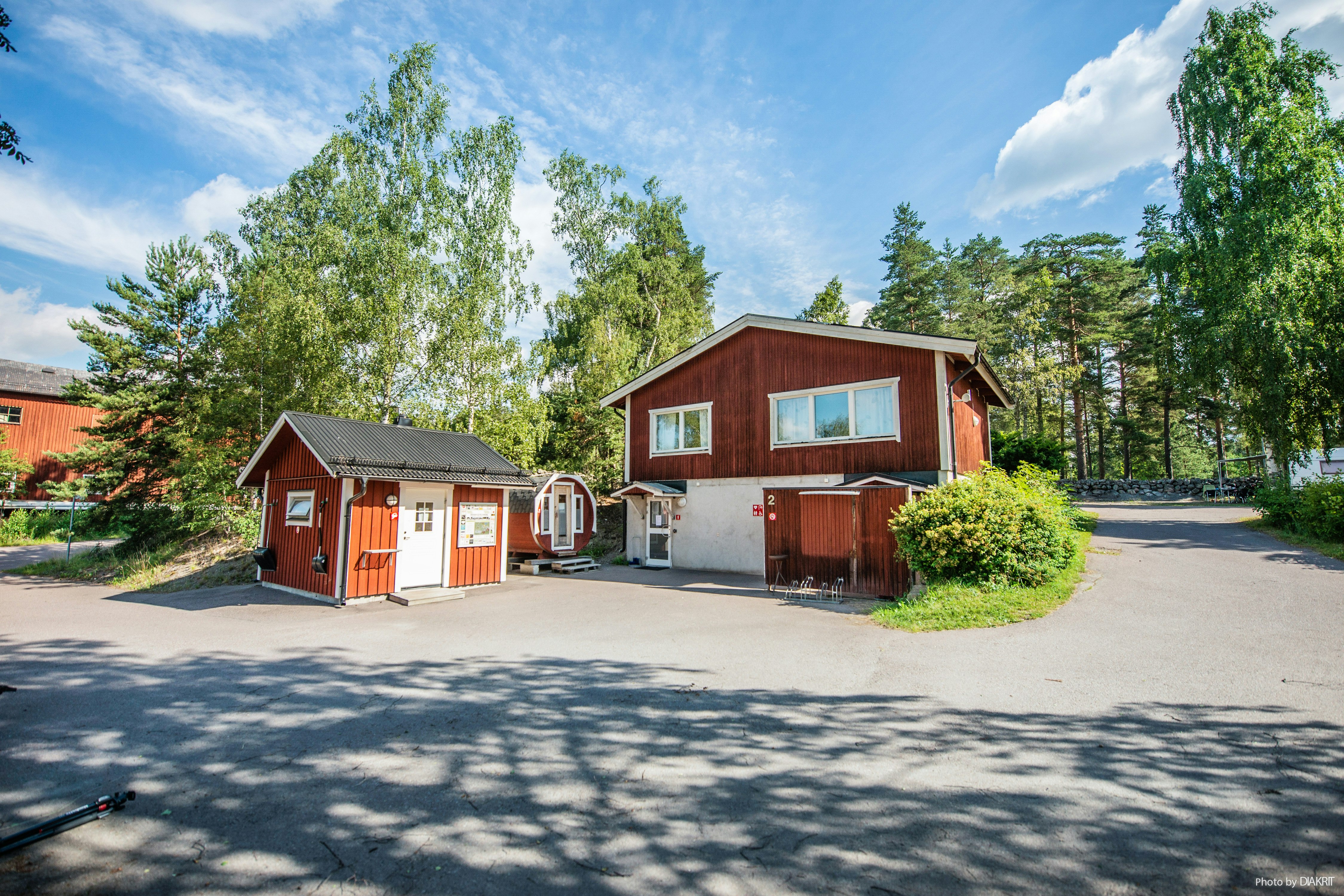 Rote Holzhäuser und ein Schuppen auf einem Campingplatz, mit Wegen, Büschen und Bäumen im Hintergrund.