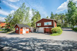 Rote Holzhäuser und ein Schuppen auf einem Campingplatz, mit Wegen, Büschen und Bäumen im Hintergrund.