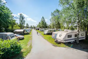 Campingplatz mit Wohnwagen und Autos entlang eines breiten Weges, umgeben von Bäumen und gepflegten Rasenflächen.