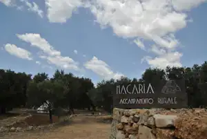Eingangsschild „Macaria“ am Campingplatz, mit Steinmauer, Kiesweg und Bäumen im Hintergrund.