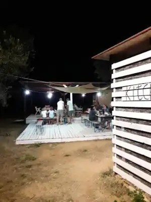 Camping-Terrasse am Abend mit Tischgruppe, Lichterkette und Holzwand, im Hintergrund Bäume und Rasenfläche.