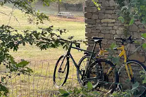 Fahrrad am Campingzaun vor einer Steinmauer, im Hintergrund Wiese und ein Tisch mit Bank.
