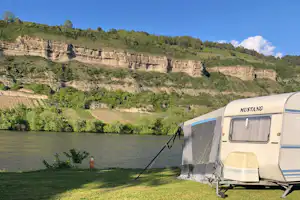Wohnwagen mit Vorzelt am Campingplatz am Fluss, im Hintergrund bewaldete Hügel und Terrassen am Ufer.