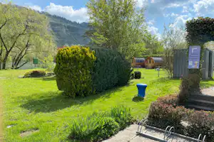 Campinggarten mit Hecken, gepflastertem Weg und Rasenfläche, im Hintergrund ein Wohnwagen und Berge.