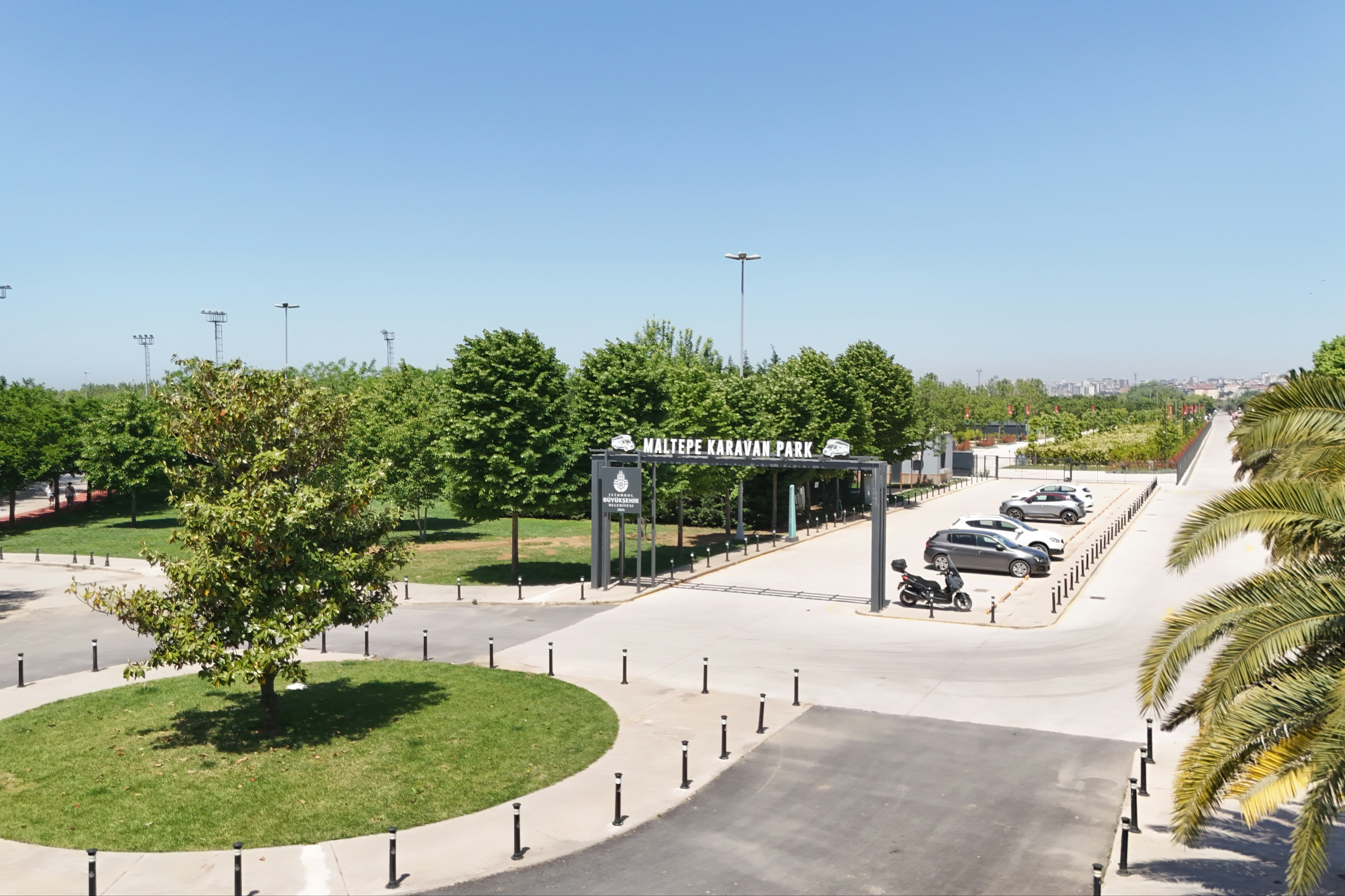 Eingang eines Campingplatzes mit Schild, breitem Weg, geparkten Autos und gepflegten Grünflächen mit Bäumen und Palmen.