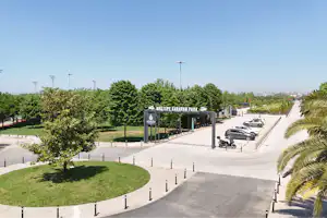 Eingang eines Campingplatzes mit Schild, breitem Weg, geparkten Autos und gepflegten Grünflächen mit Bäumen und Palmen.