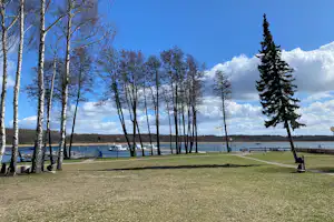 Grüner Campingplatz mit Rasenfläche, schmalem Weg und hohen Bäumen am Seeufer, im Hintergrund Wasser und Himmel.