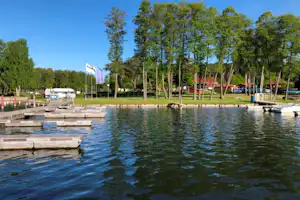 See mit Holzstegen und Liegeplätzen am Campingplatz, im Hintergrund Bäume und ein Bootssteg mit Booten.