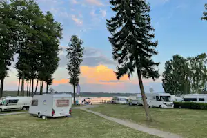 Wohnwagen auf einer Campingwiese am See, mit Bäumen und einem Kiesweg zwischen den Stellplätzen.