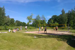 Volleyballfeld am Campingplatz: gelbes Netz über Sand, Spieler am Rand und Rasenfläche mit Bäumen im Hintergrund.