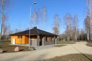 Campinghaus mit Terrasse, Kiesweg und Straßenlaterne vor einer Reihe kahler Bäume, im Hintergrund ein Parkplatz.