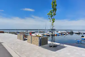 Hafenpromenade mit gepflastertem Weg, Steinmauer und jungen Bäumen; im Hintergrund liegen Segelyachten am Wasser.
