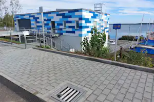Blau-weißes Campinggebäude mit Treppenanlage, gepflastertem Weg und Absperrung, im Hintergrund Segelboote am Hafen.