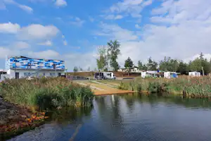 Campingplatz am Fluss mit Wohnwagen, Ufergras und einem Gebäude im Hintergrund, daneben ein Weg und Stellplätze.