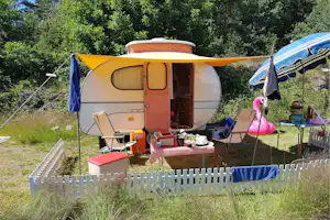 Wohnwagen mit geöffnetem Eingang, gelbem Vordach und Campingmöbeln auf dem Stellplatz, im Grünen mit Hecke und Zaun.