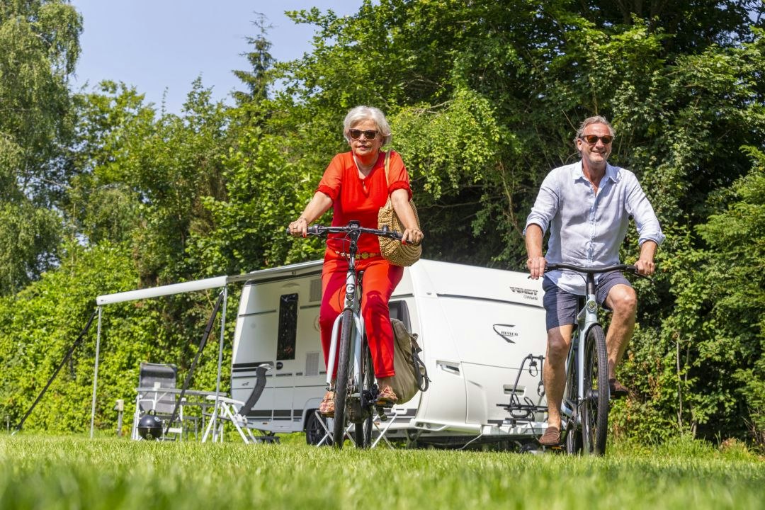 Zwei Personen fahren mit Fahrrädern vor einem Wohnwagen im Grünen, mit Campingstühlen und Tisch am Stellplatz.