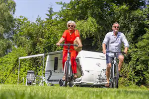 Zwei Personen fahren mit Fahrrädern vor einem Wohnwagen im Grünen, mit Campingstühlen und Tisch am Stellplatz.