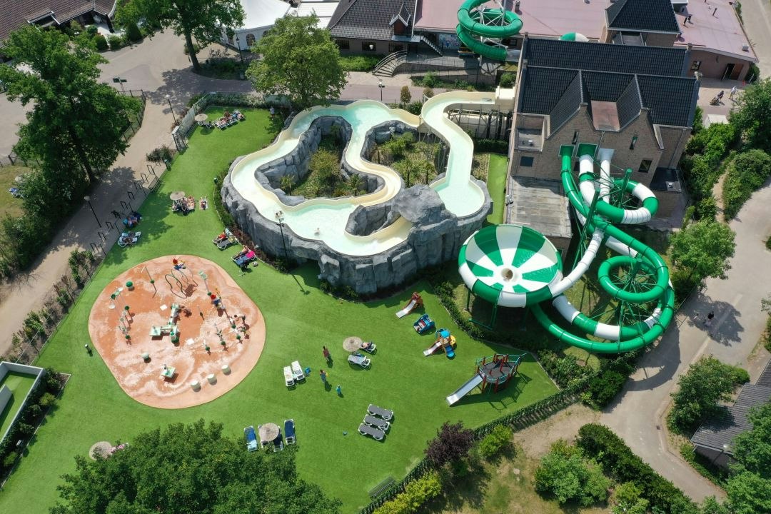Luftaufnahme eines Campingplatzes mit Wasserrutsche, Planschbecken und Spielbereich auf einer grünen Wiese zwischen Häusern.