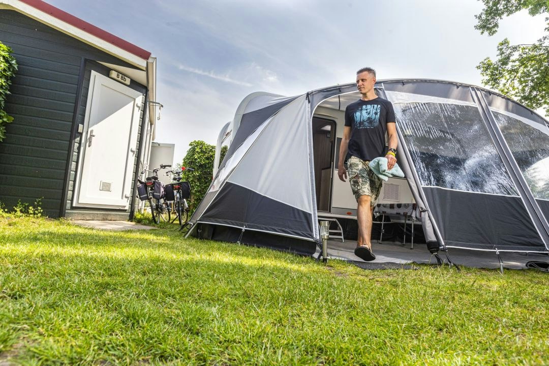 Zelt am Campingplatz neben einem Wohnwagen, Mann trägt eine Tasche über den Rasen, Fahrrad und Gebäude im Hintergrund.