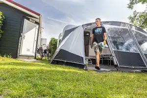 Zelt am Campingplatz neben einem Wohnwagen, Mann trägt eine Tasche über den Rasen, Fahrrad und Gebäude im Hintergrund.