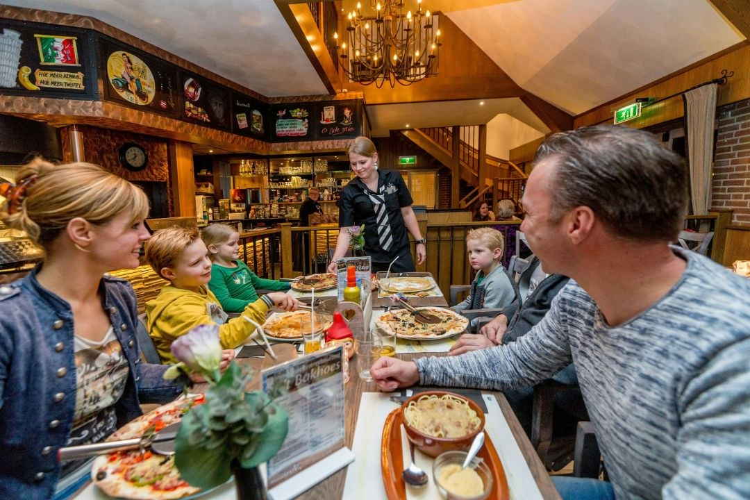 Familie sitzt an einer langen Tafel in einer Campinghütte, Kinder und Erwachsene essen, im Hintergrund eine Bar mit Getränken.