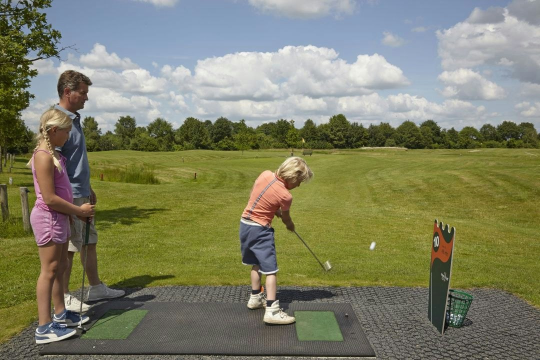 Zwei Personen stehen auf einem Golf-Übungsplatz mit Abschlagmatte, Schläger und Ball, im Hintergrund grünes Gelände und Bäume.