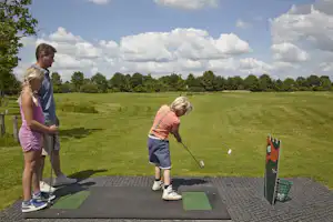 Zwei Personen stehen auf einem Golf-Übungsplatz mit Abschlagmatte, Schläger und Ball, im Hintergrund grünes Gelände und Bäume.