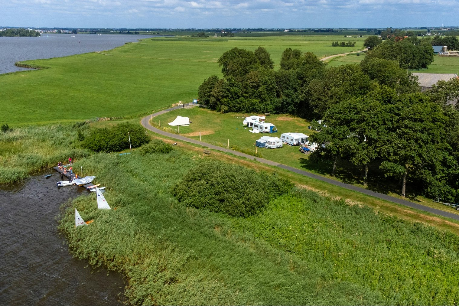 Luftaufnahme eines Campingplatzes am Fluss mit Wohnwagen, Zelten und Wegen durch grünes Gras, Bäume und Wasser im Hintergrund.
