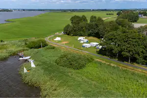 Luftaufnahme eines Campingplatzes am Fluss mit Wohnwagen, Zelten und Wegen durch grünes Gras, Bäume und Wasser im Hintergrund.