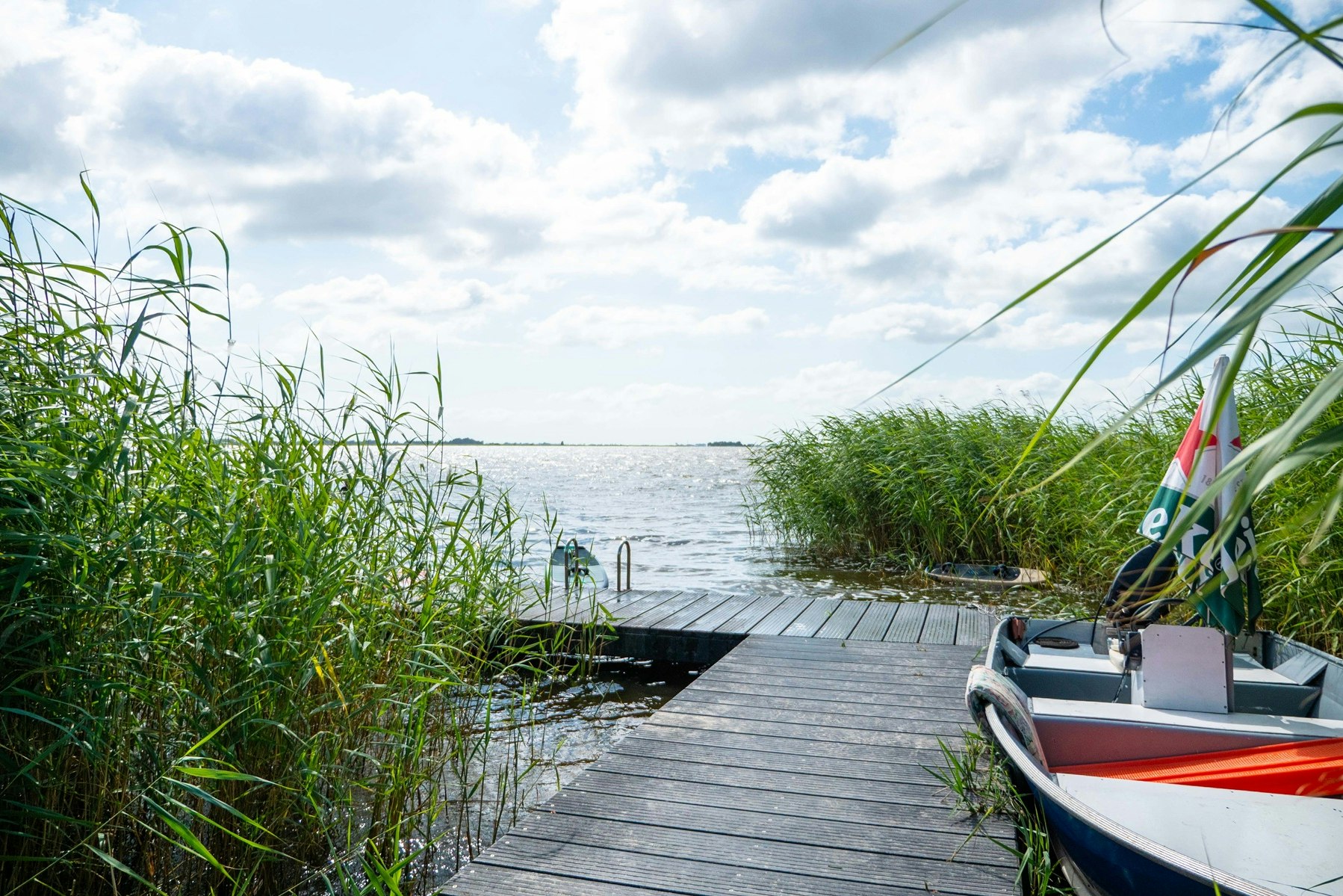 Boot am Steg am See, umgeben von Schilf und Gras, mit Blick auf das Wasser und den Himmel.