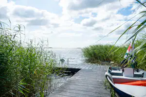 Boot am Steg am See, umgeben von Schilf und Gras, mit Blick auf das Wasser und den Himmel.