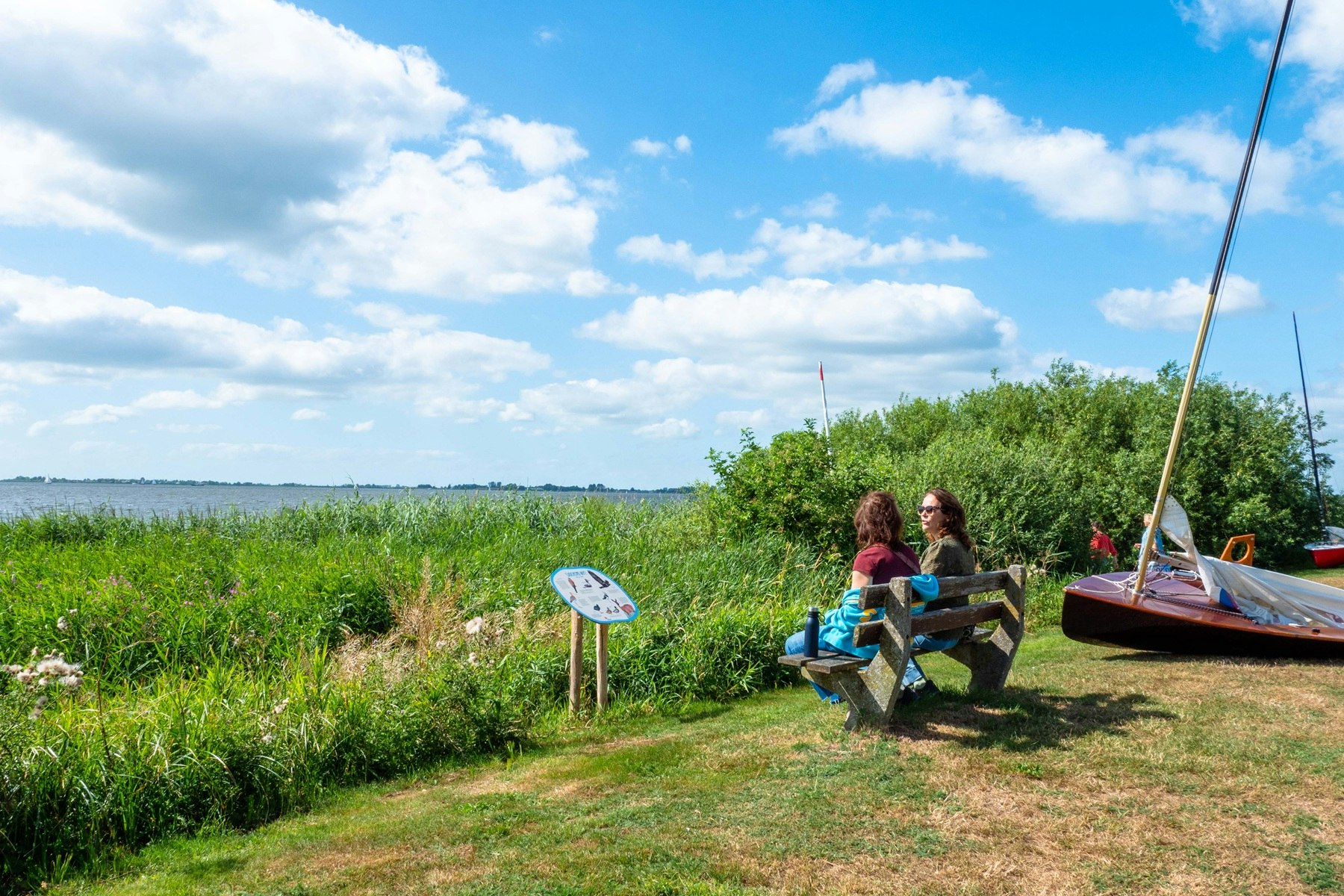 Zwei Personen sitzen auf einer Holzbank am Campingplatz, im Hintergrund Wiese, Sträucher und ein offener Himmel.