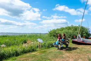 Zwei Personen sitzen auf einer Holzbank am Campingplatz, im Hintergrund Wiese, Sträucher und ein offener Himmel.