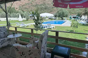 Blick von der Terrasse auf einen Campingplatz mit blauem Pool, Liegestühlen, Sonnenschirmen und grüner Wiese im Tal.