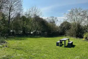 Grüner Campingplatz mit Rasenfläche, Hecken und Bäumen, im Vordergrund ein schwarzer Grill und Sitzbank.