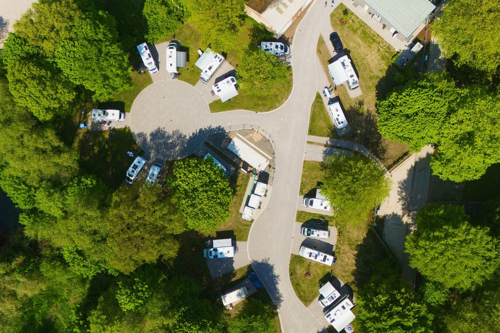 Luftaufnahme eines Campingplatzes mit mehreren Stellplätzen, Wohnwagen und Zelten, umgeben von Bäumen und einem breiten Weg.