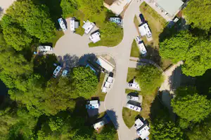 Luftaufnahme eines Campingplatzes mit mehreren Stellplätzen, Wohnwagen und Zelten, umgeben von Bäumen und einem breiten Weg.