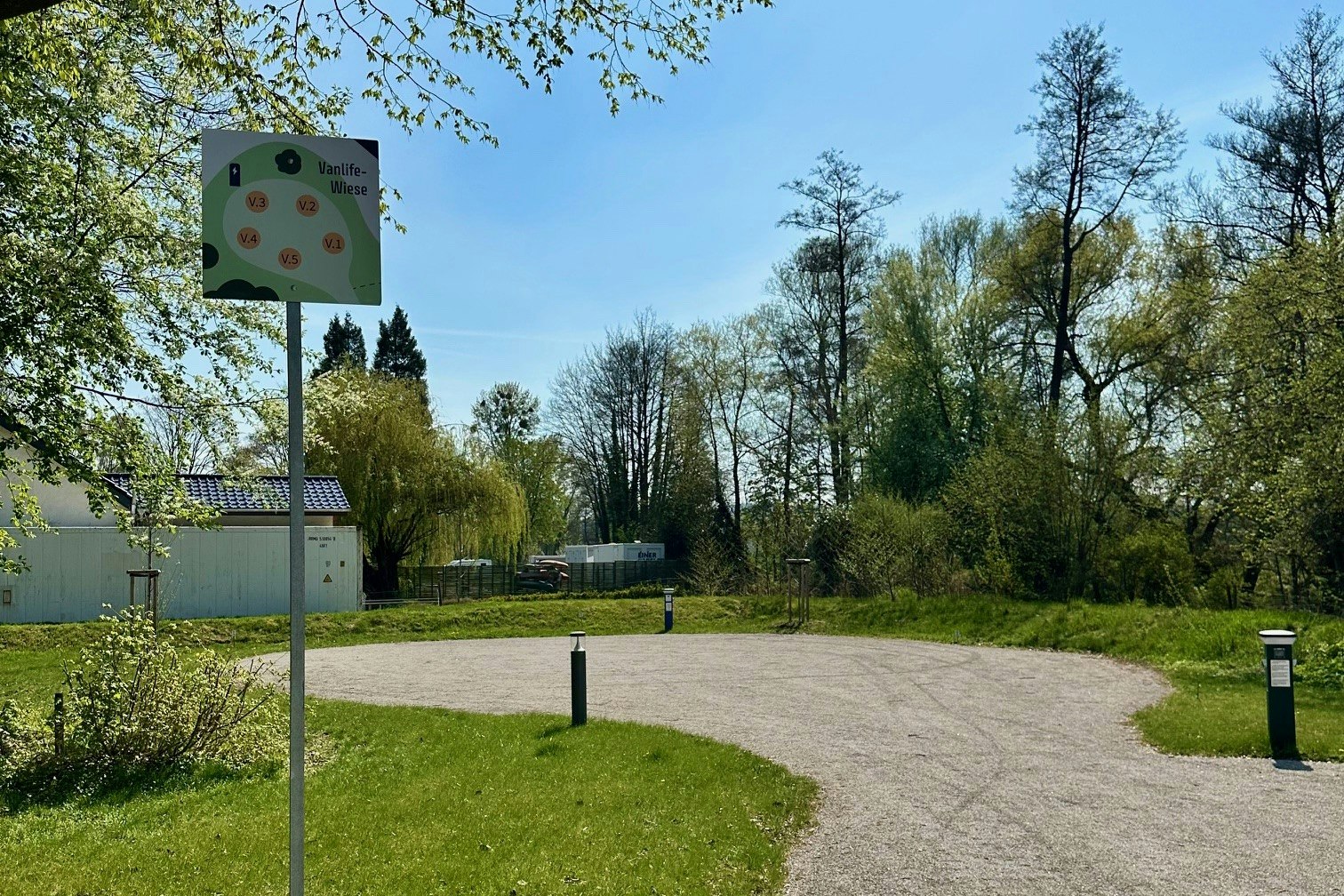 Schotterweg und Stellplatz auf einem Campinggelände, mit Wegweiser-Schild, Bäumen und Hecken im Hintergrund.