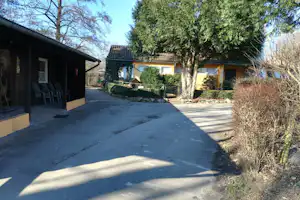 Betonierter Weg zwischen Ferienhäusern, links überdachte Terrasse mit Holzmöbeln, rechts Hecken und Bäume im Campingbereich.