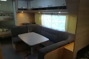 Eck-Sitzgruppe mit weißem Tisch in einem Camping-Mobilhome, daneben Vorhänge und Holzverkleidung, Schrankfächer im Hintergrund.