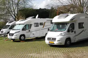 Wohnmobile auf einem gepflasterten Campingplatz, mehrere Fahrzeuge nebeneinander vor Bäumen und Hecken.