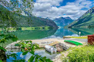 Campingplatz am See mit Blick auf Berge, Steg und Wohnwagen am Ufer, umgeben von Bäumen und Kiesweg.