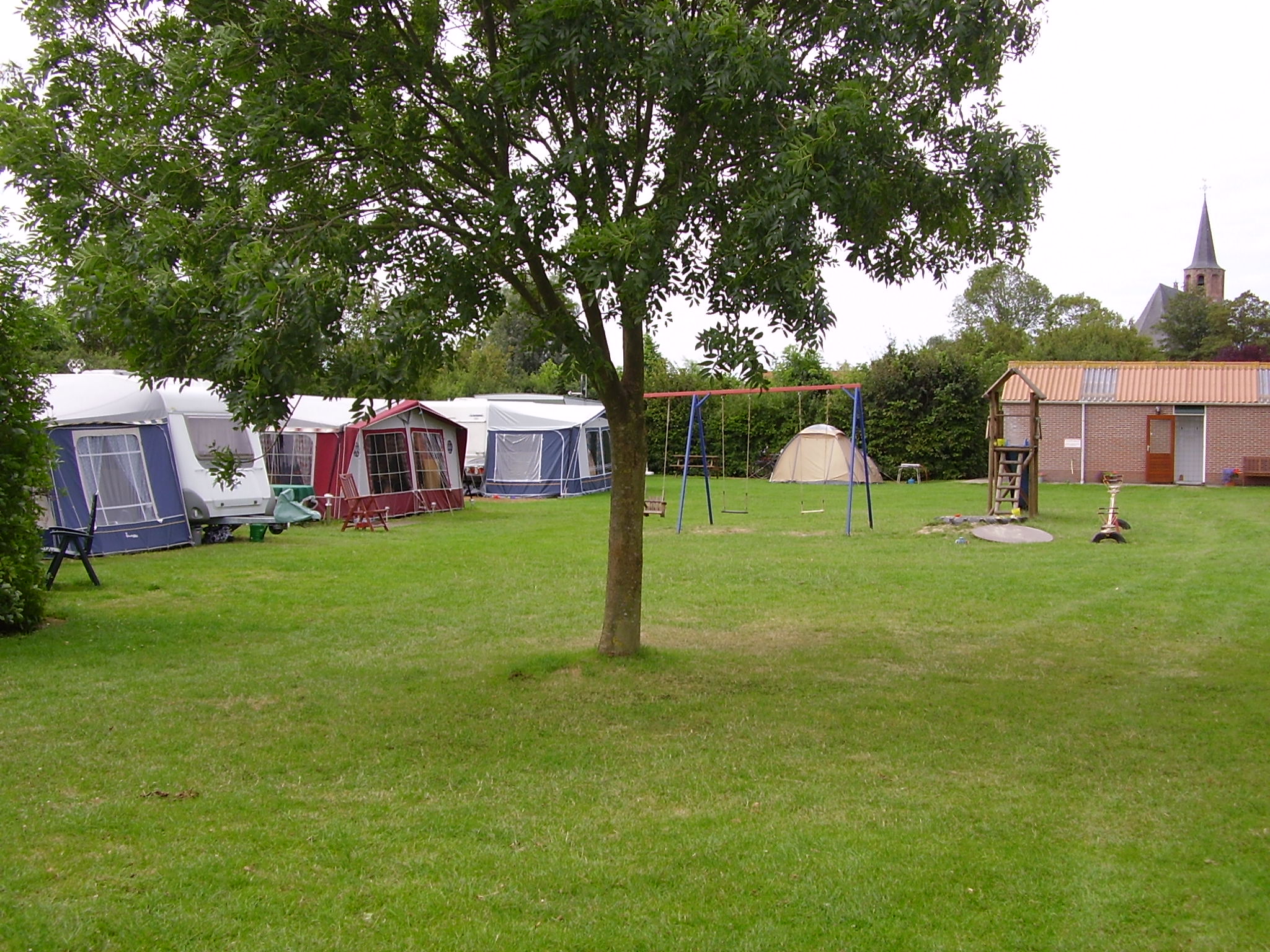 Campingwiese mit mehreren Zelten, Spielgeräten und einem Baum im Vordergrund, im Hintergrund Gebäude und Kirchturm.