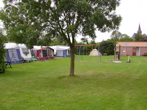 Campingwiese mit mehreren Zelten, Spielgeräten und einem Baum im Vordergrund, im Hintergrund Gebäude und Kirchturm.