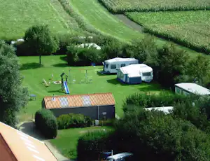 Campingplatz auf einer grünen Wiese mit mehreren Wohnwagen und Zelten, Hecken, Bäumen und einem kleinen Gebäude im Hintergrund.